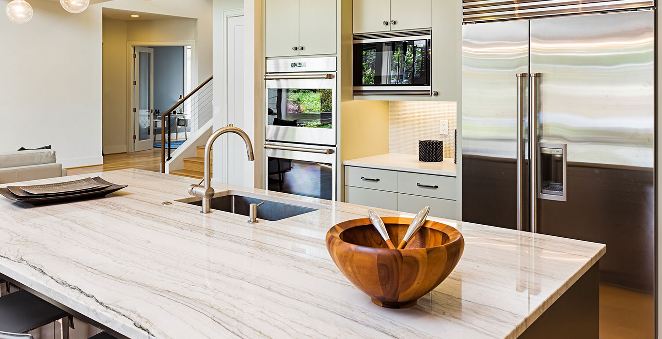 A marble kitchen countertop with a wooden bowl, silver faucet, and a fridge